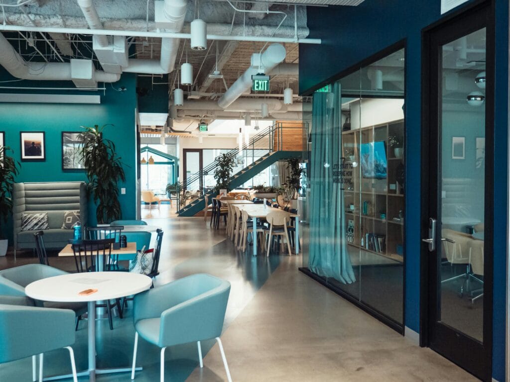 A modern coworking space with an open layout, featuring various seating options, including round tables with chairs, a high-backed booth, and long communal desks.