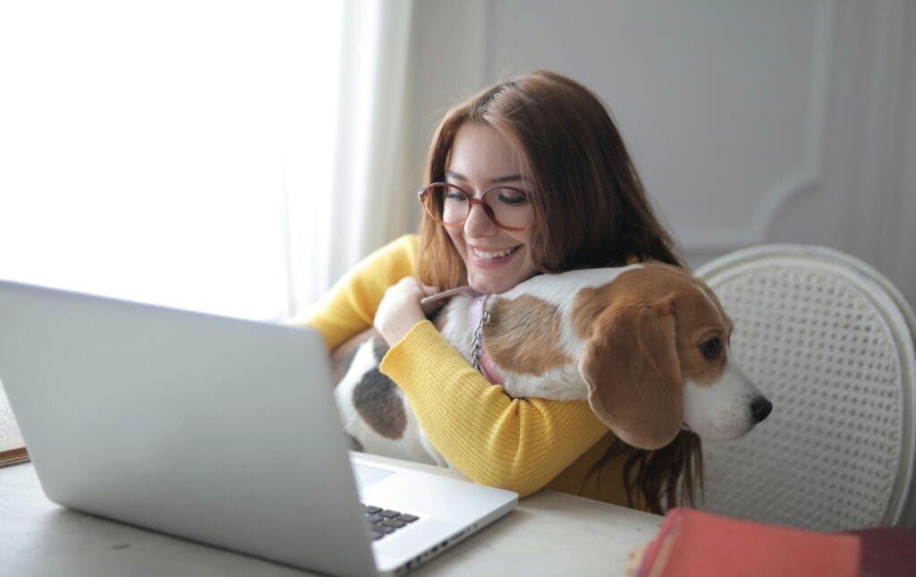 Smiling woman in glasses hugging her dog while working on a laptop in a pet-friendly workspace